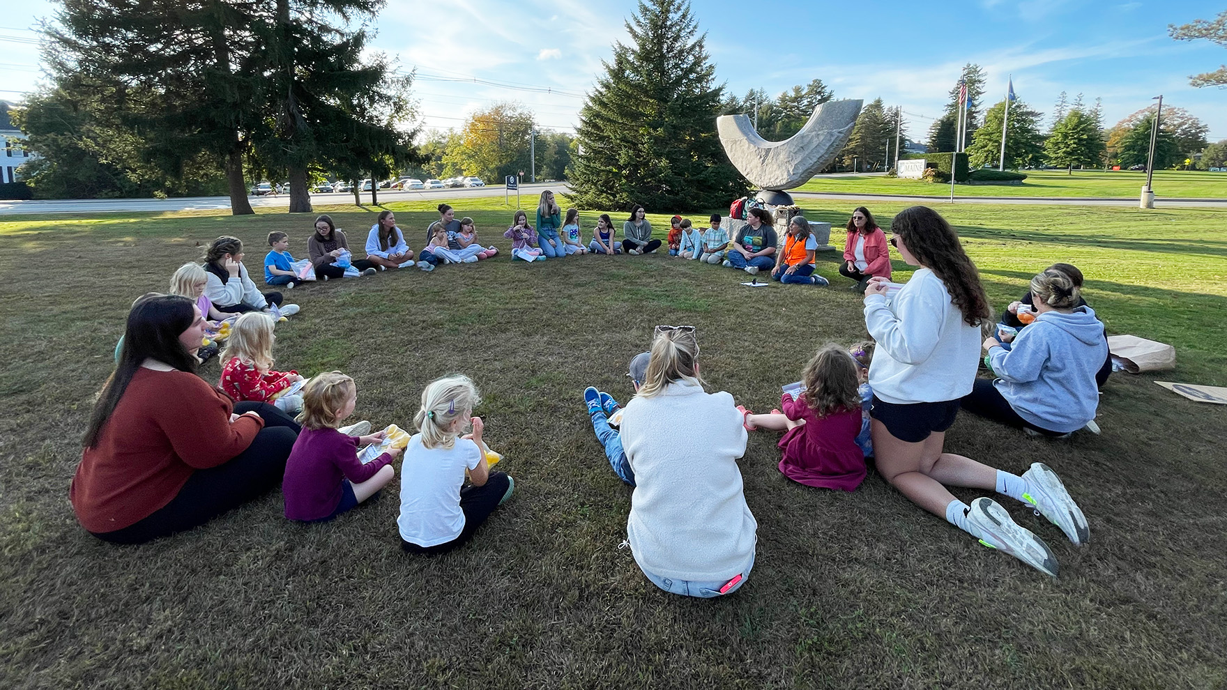 An outdoor photo of University of Maine students sitting in a circle with preschool students.